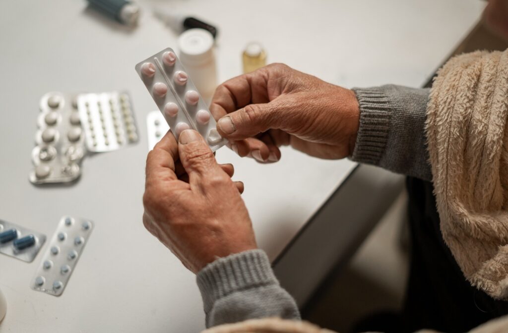 a close-up of an elderly person's hands holding a medicine packet with other prescriptions in the background on a table.