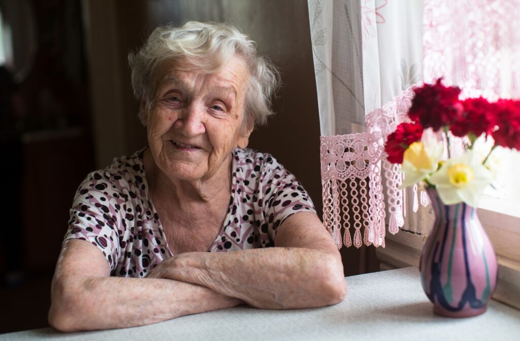 An older adult smiles and sits with their arms crossed at a small kitchen table with a vase of colorful flowers